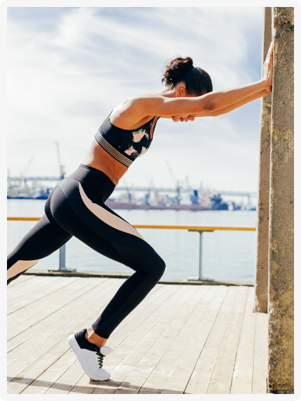 Sportos nő nyújtó mozdulat közben erősen a falnak támaszkodik, koncentrált testtartásban a vízparton.