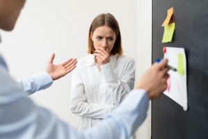 Egy nő, aki szorongva figyeli, ahogy valaki mutat neki valamit a táblán. Az arcán aggodalom és stressz tükröződik, miközben egy beszélgetés közepén van.