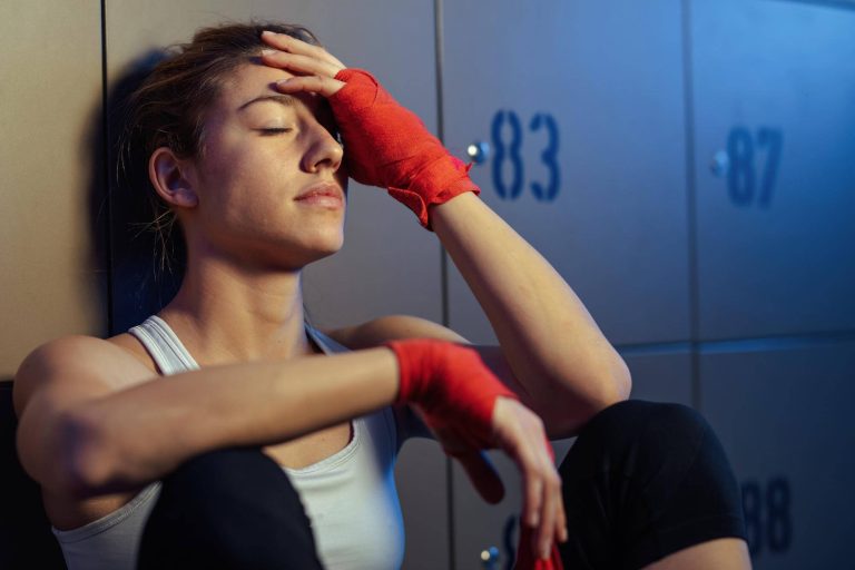 Fiatal sportoló nő ül az öltözőben, lehunyt szemmel, homlokát érintve, verseny előtti feszültséget és testi terhelést megélve.